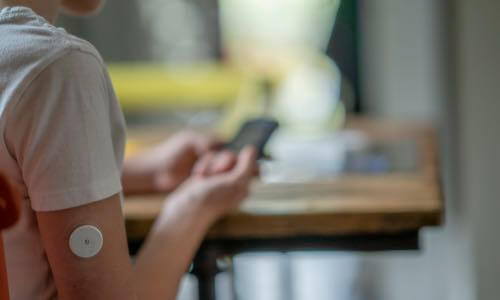 Close-up of right profile of young person at desk with CGM sensor on arm and monitor in hand