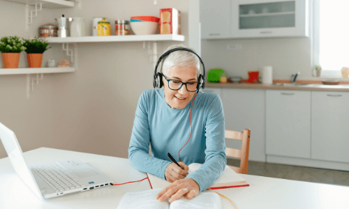 Middle-aged woman sitting at a kitchen table with open laptop, book, pen & paper while talking on a headset