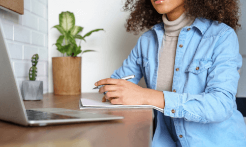 Young diverse female sitting at a counter looking at her laptop with paper & pen in hand