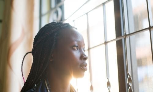 Young diverse woman sitting on a bed & looking out the window through an iron headboard