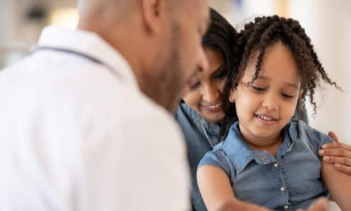 Diverse male health care professional assisting a smiling diverse female child as her mother holds her child as her mother holds her