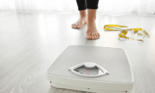 Close-up of adult bare feet facing a scale with measuring tape on the floor beside it