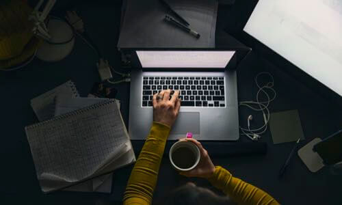 Aerial view of laptop on a desk with someone's left hand typing & right hand holding a coffee cup