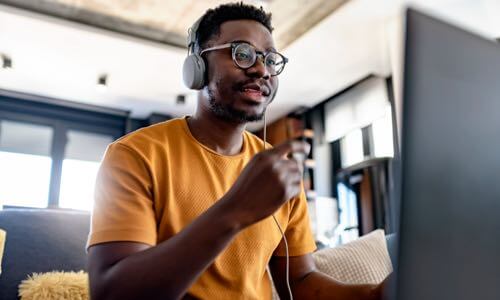 Diverse adult male sitting on a sofa, wearing a headset & speaking to someone on his laptop monitor