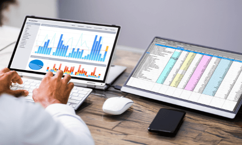 Close-up of hands typing on a white keyboard & viewing data on two hybrid laptop displays