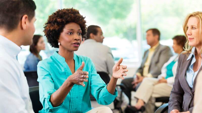 Diverse young woman sitting in a room full of professionals & speaking to a small group of people